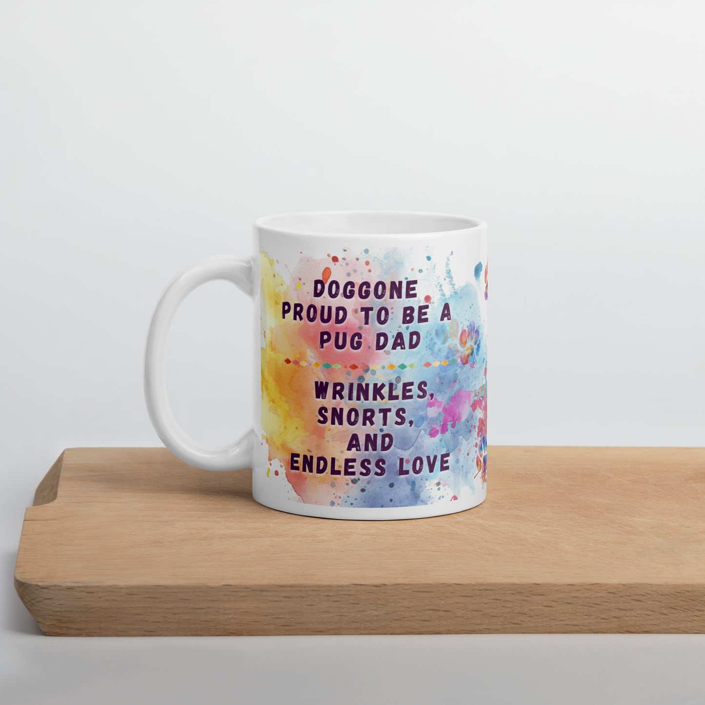 Doggone Proud Pug Dog Dad Mug. Mug with handle on left showing a cute quote: Doggone Proud to be a Pug Dad, Wrinkles, Snorts, and Endless Love. The quote is surrounded by colorful and fun watercolor splashes & paws. On a wood board.