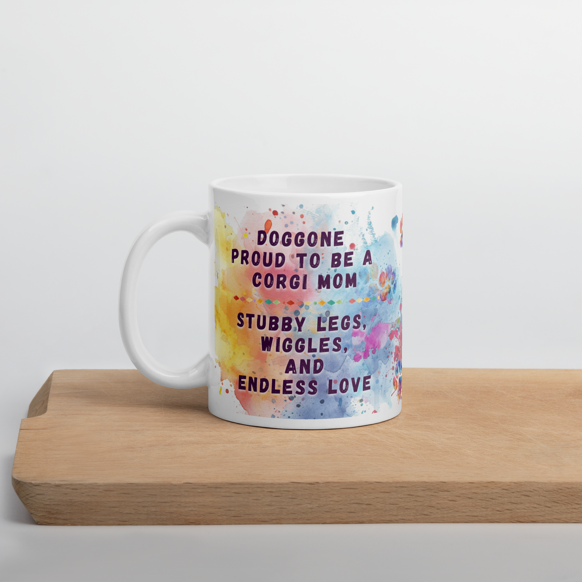Doggone Proud Corgi Dog Mom Mug. Mug with handle on left showing a cute quote: Doggone Proud to be a Corgi Mom, Stubby Legs, Wiggles, and Endless Love. The quote is surrounded by colorful and fun watercolor splashes & paws. On a wood board.