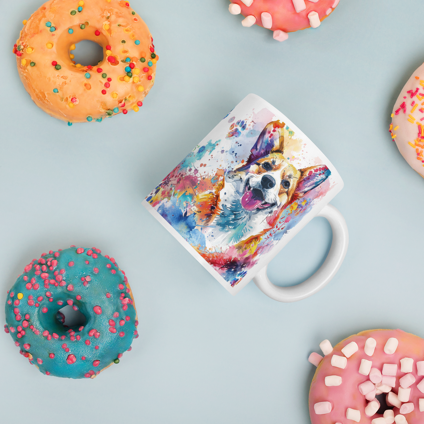 Doggone Proud Corgi Dog Mug. A fun colorful mug with handle showing a happy Corgi dog surrounded by watercolor splashes, clouds, and paws. Mug is laying flat with donuts around it.