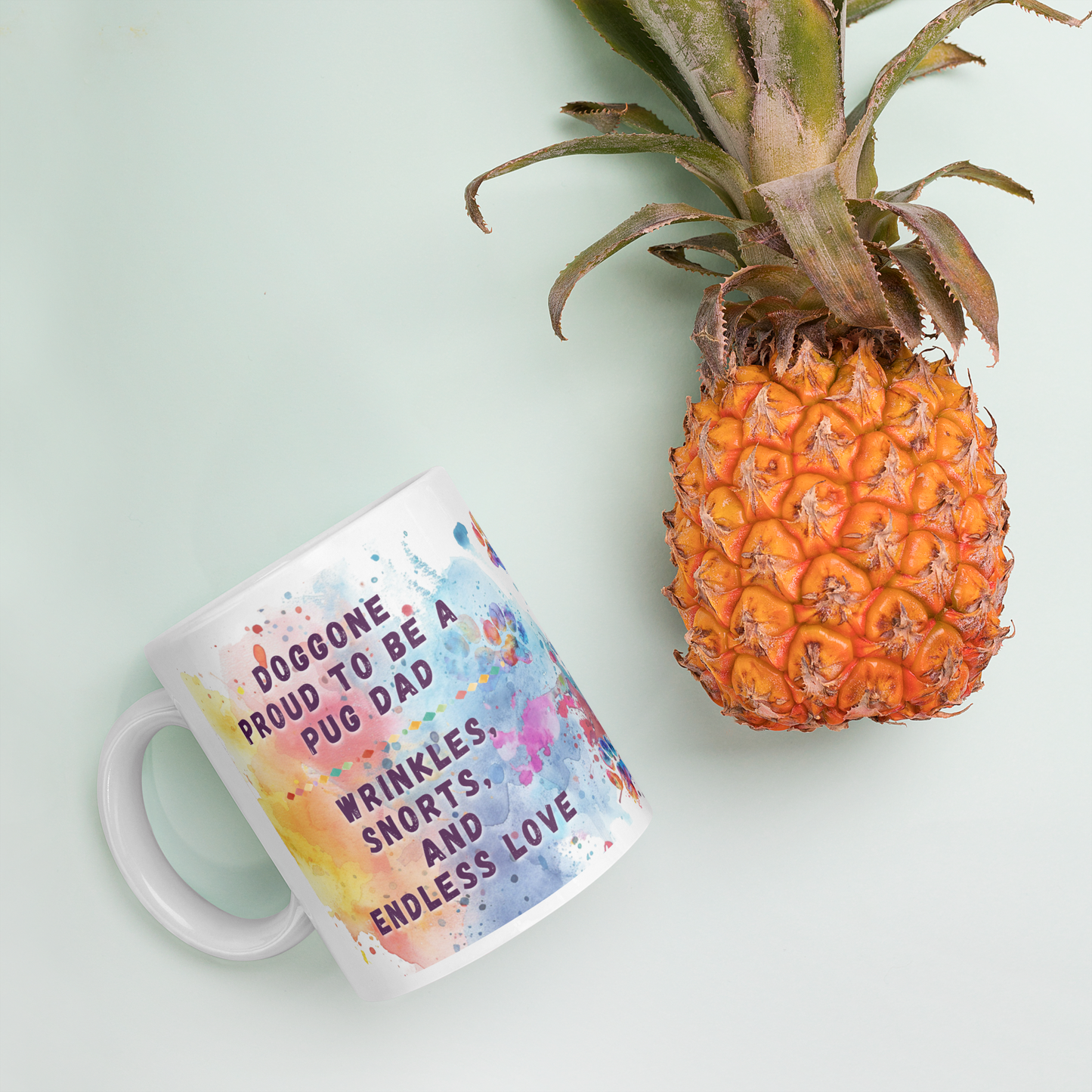 Doggone Proud Pug Dog Mug. Mug with handle on left showing a cute quote: Doggone Proud to be a Pug Dad, Wrinkles, Snorts, and Endless Love. The quote is surrounded by colorful and fun watercolor splashes & paws. Mug is laying flat with a pineapple.