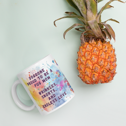 Doggone Proud Pug Dog Mug. Mug with handle on left showing a cute quote: Doggone Proud to be a Pug Dad, Wrinkles, Snorts, and Endless Love. The quote is surrounded by colorful and fun watercolor splashes & paws. Mug is laying flat with a pineapple.