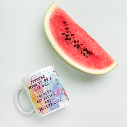 Doggone Proud Lab Dog Mug. Mug with handle on left showing a cute quote: Doggone Proud to be a Lab Dad, Loyalty, Wet Kisses, and Endless Love. The quote is surrounded by colorful and fun watercolor splashes & paws. Mug is laying flat with a watermelon.