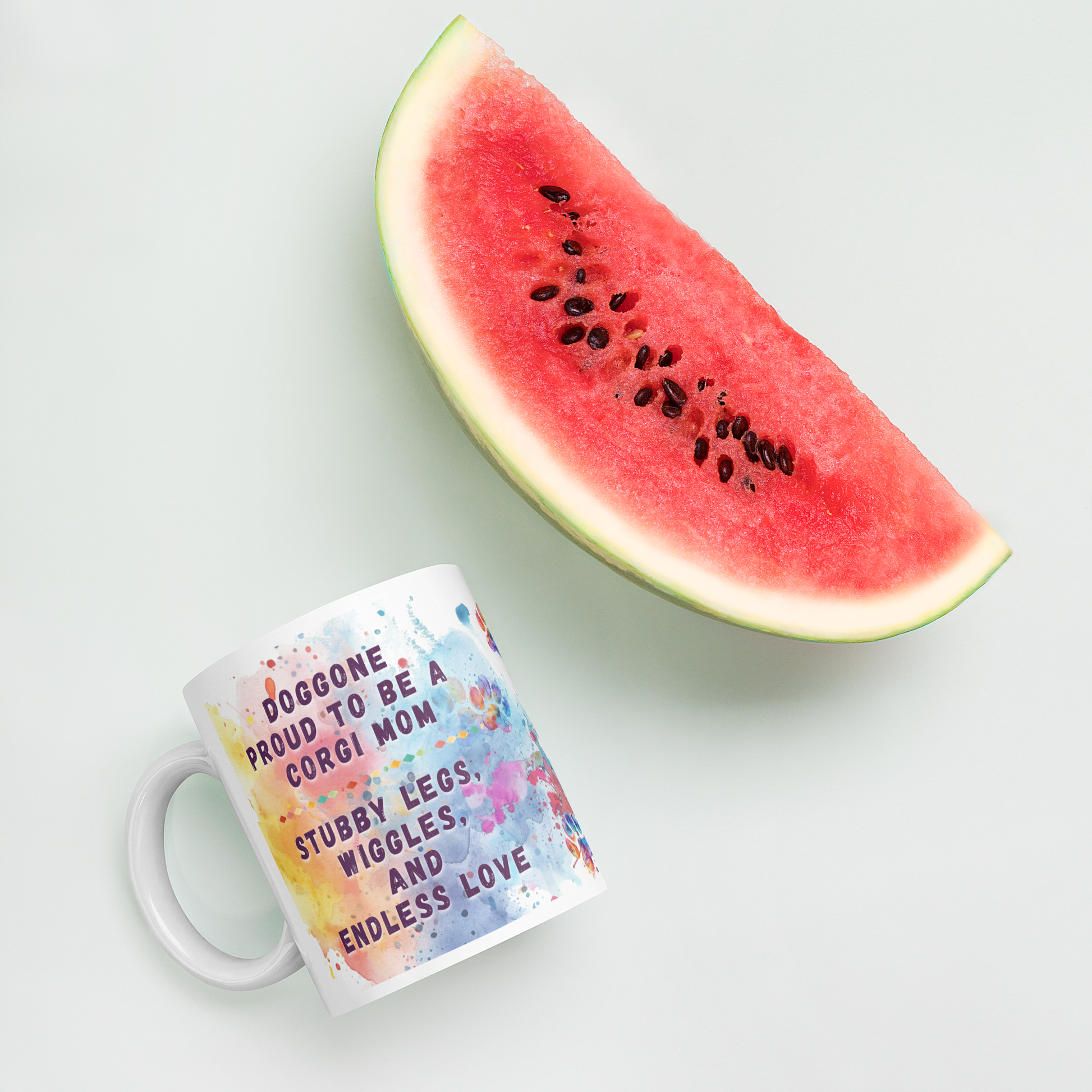 Doggone Proud Corgi Mug. Mug with handle on left showing a cute quote: Doggone Proud to be a Corgi Mom, Loyalty, Wet Kisses, and Endless Love. The quote is surrounded by colorful and fun watercolor splashes & paws. Mug is laying flat with a watermelon.