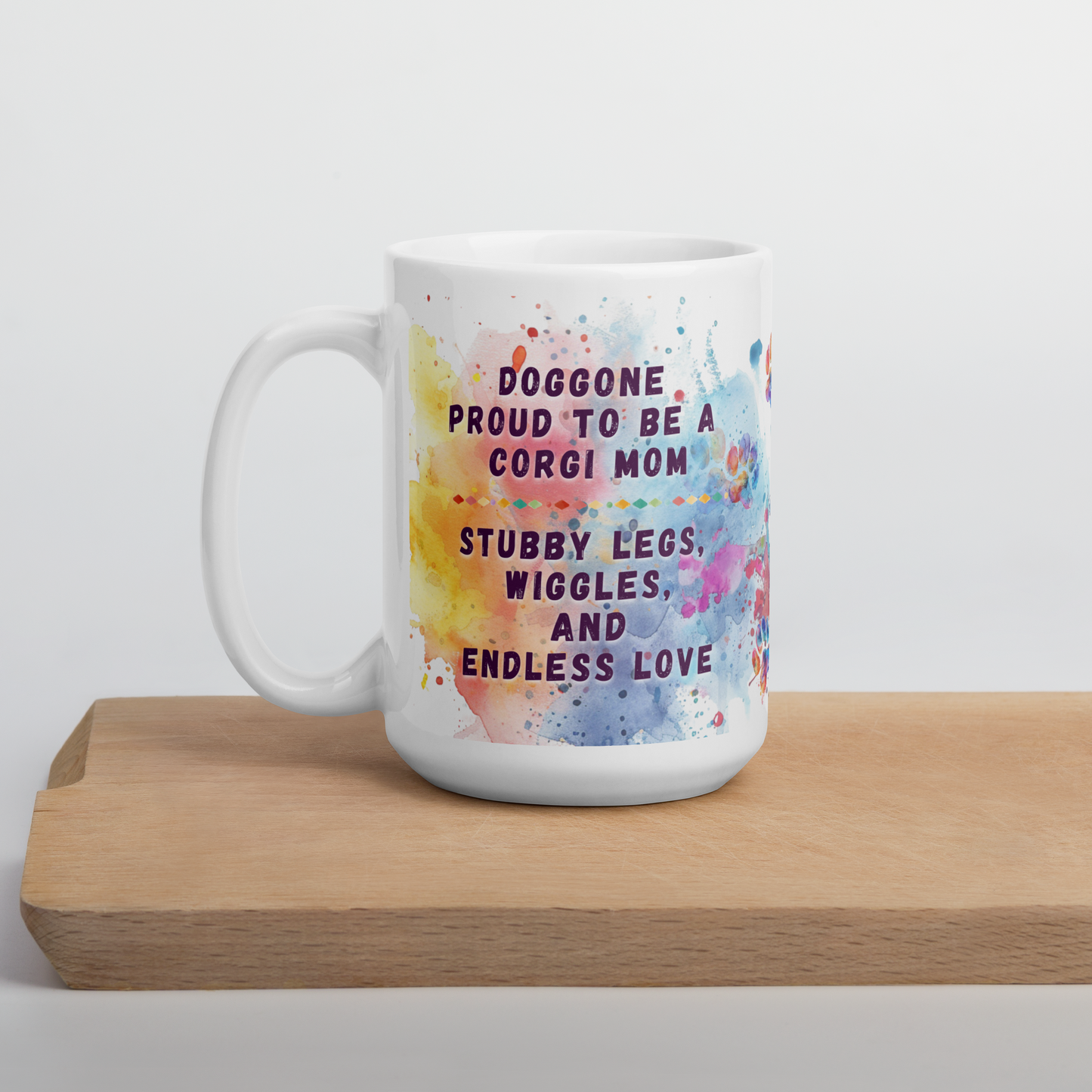Doggone Proud Corgi Dog Mom Mug. Mug with handle on left showing a cute quote: Doggone Proud to be a Corgi Mom, Stubby Legs, Wiggles, and Endless Love. The quote is surrounded by colorful and fun watercolor splashes & paws. On a wood board.