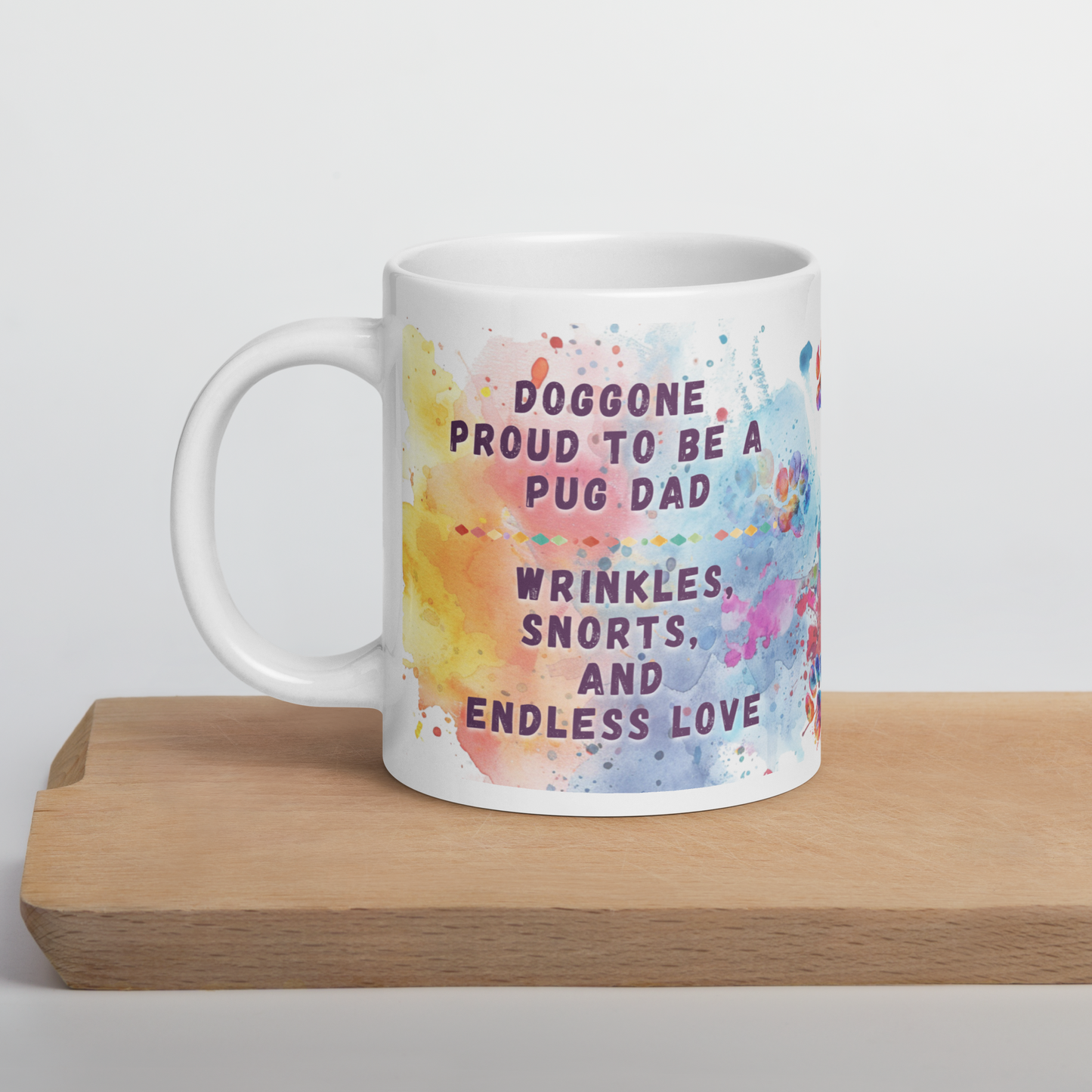 Doggone Proud Pug Dog Dad Mug. Mug with handle on left showing a cute quote: Doggone Proud to be a Pug Dad, Wrinkles, Snorts, and Endless Love. The quote is surrounded by colorful and fun watercolor splashes & paws. On a wood board.