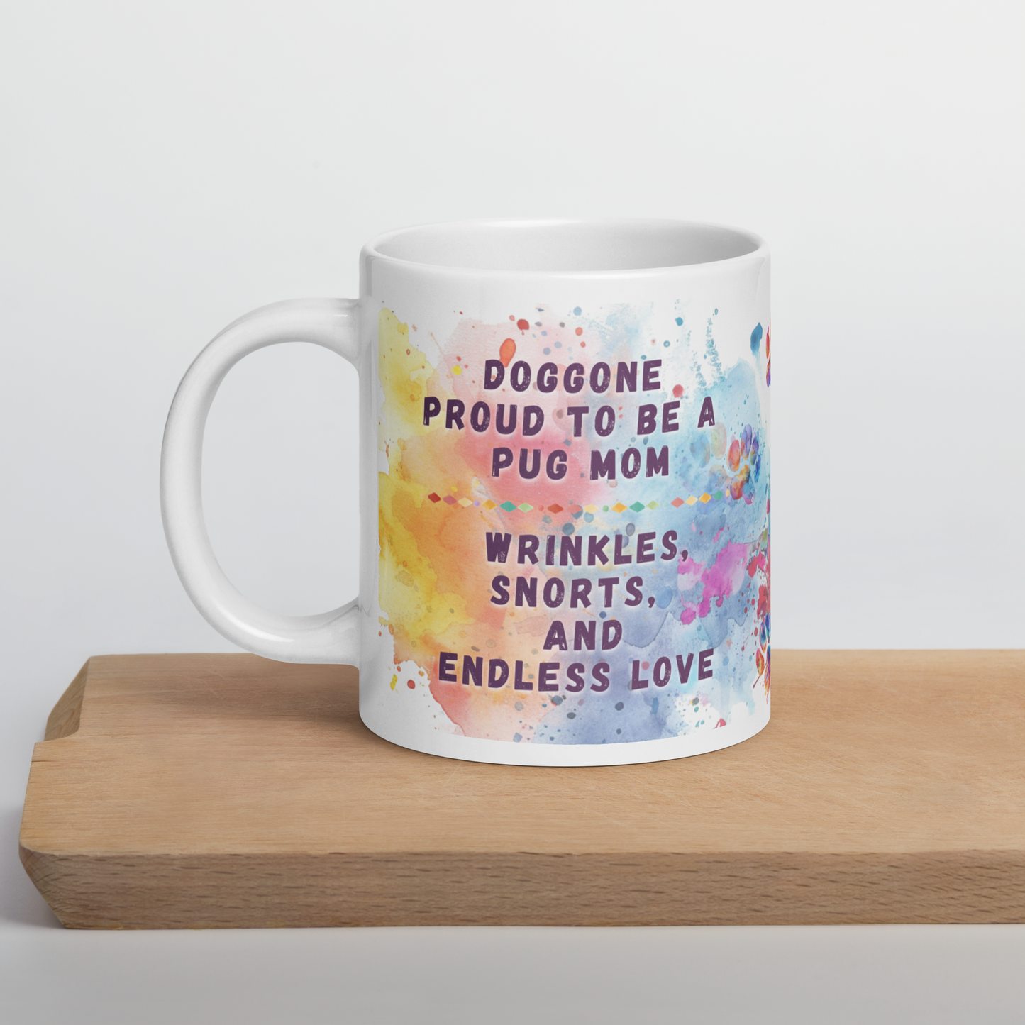 Doggone Proud Pug Dog Mom Mug. Mug with handle on left showing a cute quote: Doggone Proud to be a Pug Mom, Wrinkles, Snorts, and Endless Love. The quote is surrounded by colorful and fun watercolor splashes & paws. On a wood board.