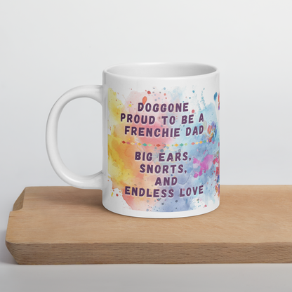 Doggone Proud Frenchie Dog Dad Mug. Mug with handle on left showing a cute quote: Doggone Proud to be a Frenchie Dad, Big Ears, Snorts, and Endless Love. The quote is surrounded by colorful and fun watercolor splashes & paws. On a wood board.