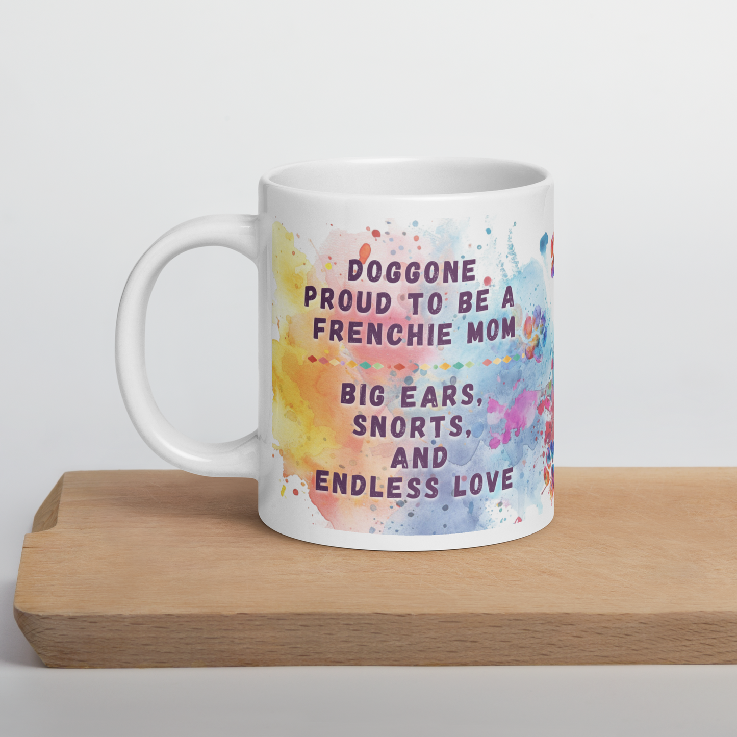 Doggone Proud Frenchie Dog Mom Mug. Mug with handle on left showing a cute quote: Doggone Proud to be a Frenchie Mom, Big Ears, Snorts, and Endless Love. The quote is surrounded by colorful and fun watercolor splashes & paws. On a wood board.