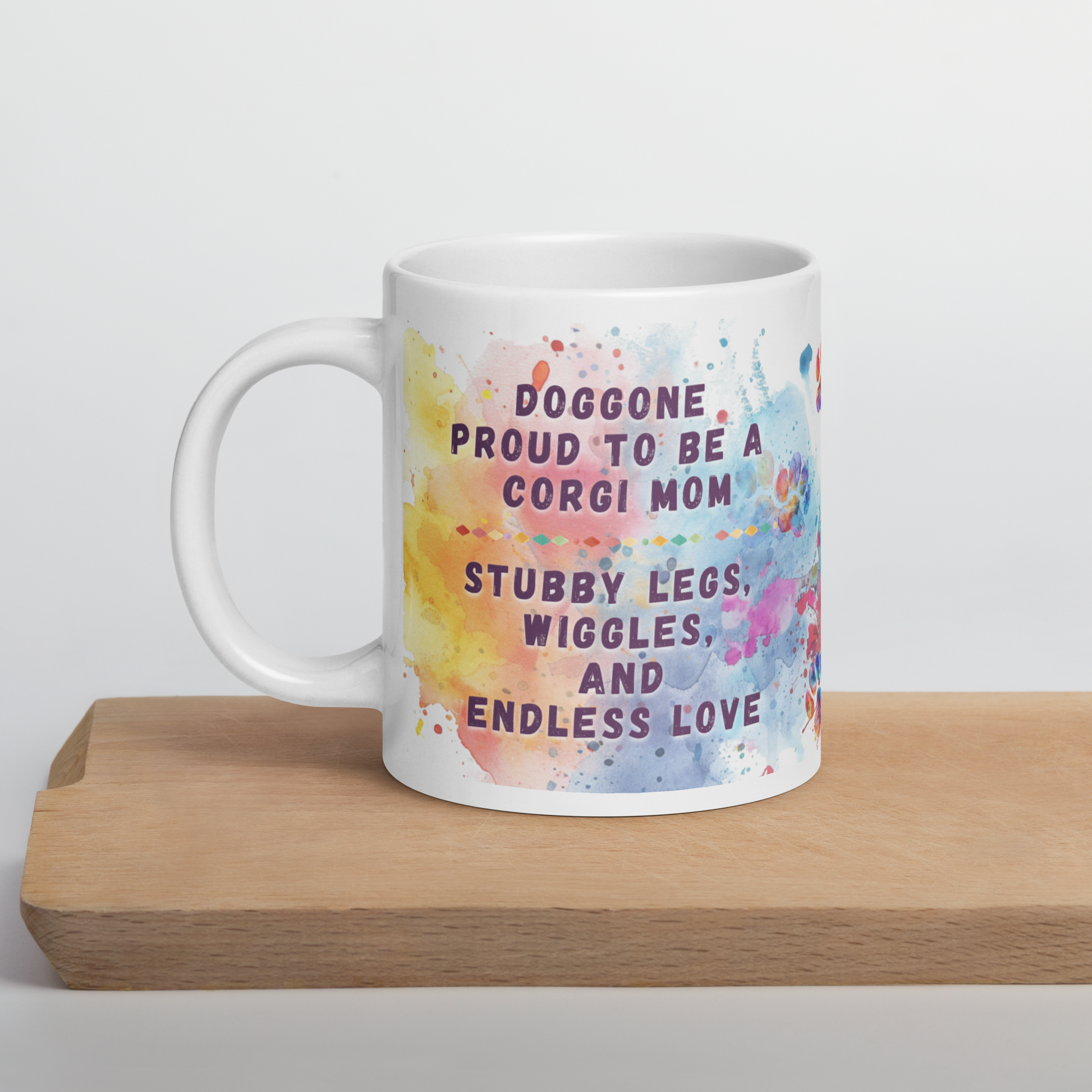 Doggone Proud Corgi Dog Mom Mug. Mug with handle on left showing a cute quote: Doggone Proud to be a Corgi Mom, Stubby Legs, Wiggles, and Endless Love. The quote is surrounded by colorful and fun watercolor splashes & paws. On a wood board.